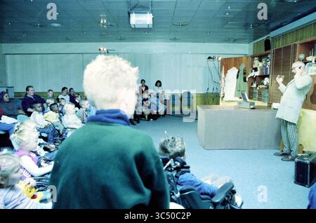 Theater für Bildung] kranke Kinder, Maria Foundation, 14-10-2003 Whizgle News, Dutch Desk, Niederlande, 1950-2000 Ein Darsteller fesselt ein fesselndes Publikum aus Kindern und Erwachsenen in einer Sitzordnung mit verschiedenen Requisiten auf einem Tisch. Die Atmosphäre lässt eine lebendige und interaktive Präsentation vermuten, die wahrscheinlich unterhaltsam oder lehrreich ist. Das Publikum, einschließlich Kinder im Rollstuhl und andere, scheint aufmerksam zu sein, während es zuhört und beobachtet. Stockfoto