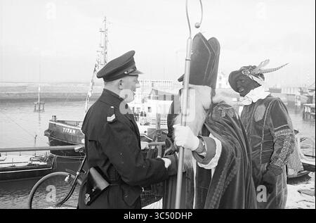 Ankunft von St. Nikolaus in IJmuiden, St. Nikolaus, Sinterklaas, 27-11-1965 Whizgle News, Dutch Desk, Niederlande, 1950-2000 Ein Polizeibeamter begrüßt eine Figur in traditioneller Bischofskleidung, begleitet von einer anderen Person in farbenfroher historischer Kleidung, in der Nähe eines Hafens mit Booten im Hintergrund. Stockfoto