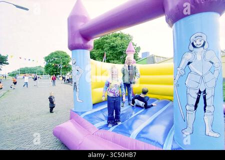 Street Play Day, Howler Buoy, IJmuiden, IJmuiden, Niederlande, 23-05-2000 Whizgle News, Dutch Desk, Niederlande, 1950-2000 In Einem lebhaften Hüpfhaus spielen Kinder fröhlich, wobei ein Kind springt, während ein anderes sich auf das Rutschen vorbereitet. Farbenfrohe Dekorationen und festliche Elemente umgeben die Szene und sorgen für eine lebhafte Atmosphäre bei einer Gemeinschaftsveranstaltung. Stockfoto