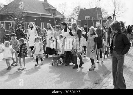 Zwaanshoek Parade, Queen's Day, 30 04.04.1976 Whizgle News, Dutch Desk, Niederlande, 1950-2000 Eine lebhafte Parade, bei der eine Gruppe von Kindern und Erwachsenen, einige in traditionellen Trachten, auf einer Straße marschieren. Einige Kinder halten die Hände, andere schieben Spielzeugwagen, während die Leute im Hintergrund die Prozession mit Lächeln beobachten. Bäume säumen die Straße und tragen zur festlichen Atmosphäre bei. Stockfoto