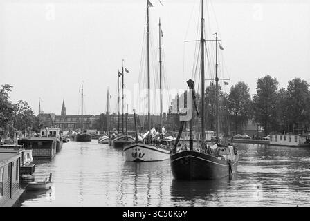 Neun Segelschiffe aus der Zeit um 1900 fahren in Haarlem an und fahren durch die Spaarne, Haarlem, Niederlande, 29-05-1990 Whizgle News, Dutch Desk, Niederlande, 1950-2000 Eine beschauliche Hafenszene mit mehreren Booten, die entlang einer ruhigen Wasserstraße vertäut sind, eingerahmt von Bäumen und Gebäuden im Hintergrund. Fahnen flattern über den Masten, während ein markanter Turm in der Entfernung zu sehen ist. Stockfoto
