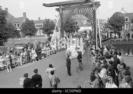 Blumenparade Aalsmeer, Blumenparade, Blumenausstellung, 04-09-1971 Whizgle News, Dutch Desk, Niederlande, 1950-2000 Eine große Menschenmenge versammelt sich entlang eines Kanals, gesäumt von Bäumen und Gebäuden, um eine festliche Prozession zu beobachten. Die Menschen zeigen eine Mischung aus Spannung und Neugier, während ein dekoriertes Fahrzeug langsam unter einer kunstvollen Brücke vorbeifährt. Uniformierte Offiziere halten Ordnung, während Kinder und Erwachsene Fotos machen und winken. Stockfoto