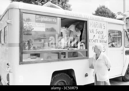 Street Trading, Belästigung Act, Market, 01-07-1982 Whizgle News, Dutch Desk, Niederlande, 1950-2000 In Einem Vintage-Fischmarkt-Wagen wird ein Händler angezeigt, der einen Kunden bedient, mit einem Schild, das für frischen Fisch steht. Die ältere Frau sieht eifrig zu, wie der Verkäufer ihre Bestellung vorbereitet und eine nostalgische Atmosphäre eines geschäftigen Straßenmarktes schafft. Stockfoto