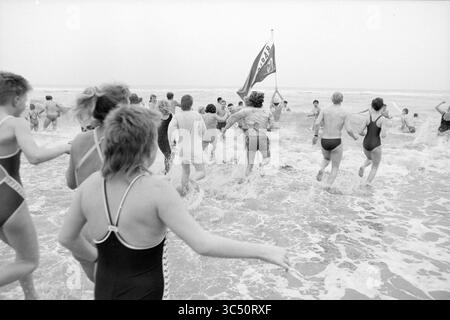 Neujahrstauchen Zandvoort, Neujahr, Zandvoort, 01-01-1987 Whizgle News, Dutch Desk, Niederlande, 1950-2000 Eine Gruppe von Menschen, die in Badeanzügen gekleidet sind, stürzt fröhlich auf die Meereswellen, während eine Flagge im Hintergrund flattert, was eine lebhafte Strandatmosphäre anzeigt. Stockfoto