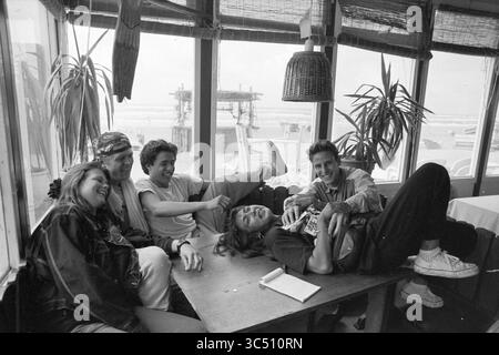 Junge Leute im Strandpavillon, Zandvoort, 10-07-1990 Whizgle News, Dutch Desk, Niederlande, 1950-2000 Eine Gruppe von fünf Freunden entspannen sich und lachen sich an einem Tisch in einem gemütlichen Café, mit Blick auf den Strand durch große Fenster. Ihre spielerischen Interaktionen und entspannten Posen fangen einen Moment der Kameradschaft und Freude ein. Stockfoto