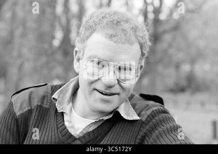 Portrait man, 00-10-1988 Whizgle News, Dutch Desk, Niederlande, 1950-2000 Ein Mann mit lockigen Haaren und Brille lächelt warm und trägt einen Pullover über einem Hemd mit Kragen. Der Hintergrund ist mit verschwommenen Bäumen versehen, die auf eine Umgebung im Freien hinweisen. Stockfoto