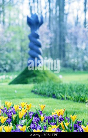 Skulpturen in einem Garten, 24-03-1999 Whizgle News, Dutch Desk, Niederlande, 1950-2000 im Vordergrund blühen gelbe und violette Tulpen, während sich im Hintergrund eine einzigartige Spiralskulptur majestätisch erhebt, umgeben von üppigem grünem Gras und Anklängen von blühenden Bäumen. Stockfoto