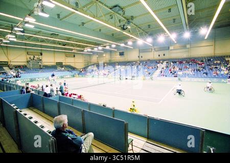 Rollstuhltennis in der Sporthalle mit Festlichkeiten in der Lobby, 00-09-1996 Whizgle News, Dutch Desk, Niederlande, 1950-2000 Eine geräumige Tennishalle bietet mehrere Spieler, die an Spielen teilnehmen, während verstreute Zuschauer einige Plätze einnehmen. Die lebhafte Oberfläche des Spielfeldes steht im Kontrast zur gedämpften Umgebung und schafft eine Atmosphäre der Vorfreude und des Wettkampfs. Stockfoto