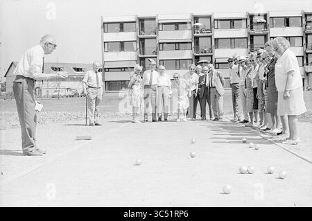 Jeu de Boules Court, 00-00-1973 Whizgle News, Dutch Desk, Niederlande, 1950-2000 Eine Gruppe von Menschen beobachtet aufmerksam, wie ein Mann sich darauf vorbereitet, einen Ball auf einem langen, schmalen Spielfeld zu Rollen, auf dem bereits mehrere Bälle auf dem Boden verstreut sind. Die Zuschauer, eine Mischung aus Männern und Frauen, zeigen eine Reihe von Ausdrucksformen, von Fokus bis Spannung, während sie in einer zwanglosen, von modernen Apartmentgebäuden umgebenen Umgebung stehen. Stockfoto