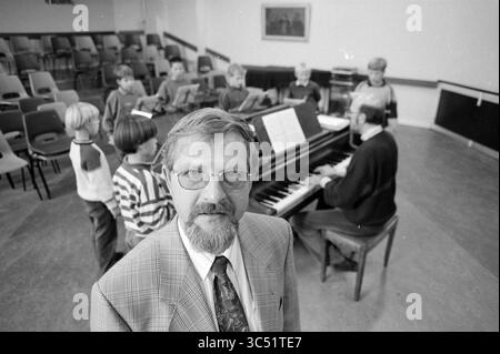 Kees besuchte 40 Jahre lang die Hörschule, 23-10-1991 Whizgle News, Dutch Desk, Niederlande, 1950-2000 Ein Mann in einem Anzug steht im Vordergrund und lächelt in die Kamera, während sich eine Gruppe von Kindern um ein Klavier im Hintergrund versammelt. Die Kinder scheinen an einem Musikunterricht beteiligt zu sein, mit Noten vor ihnen, während ein anderer Erwachsener Klavier spielt. Stockfoto