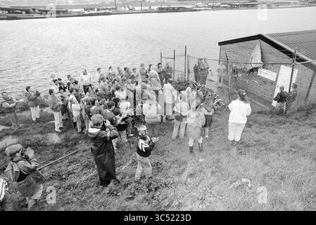 Eröffnung von Bootsschuppen, IJm., IJmuiden, Niederlande, 16-09-1994 Whizgle News, Dutch Desk, Niederlande, 1950-2000 Eine große Menschenmenge versammelt sich in der Nähe eines umzäunten Bereichs am Wasser, wobei eine Gruppe von Personen Fahrräder hält und andere aufmerksam beobachten. Die Spannung ist spürbar, wenn die Zuschauer an einem wichtigen Ereignis beteiligt sind, möglicherweise an einem Protest oder einer Gemeindeversammlung, mit einem Gebäude im Hintergrund, das ein Banner trägt. Stockfoto