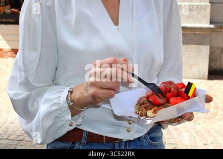 Eine Frau in der Hand einer belgischen Waffel mit Erdbeeren und Schokolade in Brügge. Street Food Szene mit belgischer Flagge und lässigem Outfit, im Freien gedreht. Stockfoto