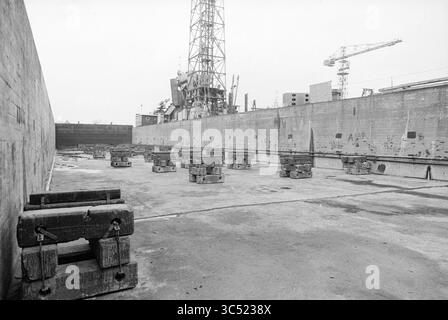 Werft Stapel Spaarndam, Spaarndam, Schiffe, 15-11-1978 Whizgle News, Dutch Desk, Niederlande, 1950-2000 eine Industrieszene mit einem großen, leeren Hafenbereich, der von Betonmauern umgeben ist. Mehrere schwere Blöcke sind ordentlich auf dem Boden angeordnet, mit Kränen und einem großen Bauwerk im Hintergrund, was auf laufende Bau- oder Reparaturarbeiten in der Nähe hindeutet. Die Atmosphäre ist schlicht und zweckmäßig, was die Größe des Raumes unterstreicht. Stockfoto