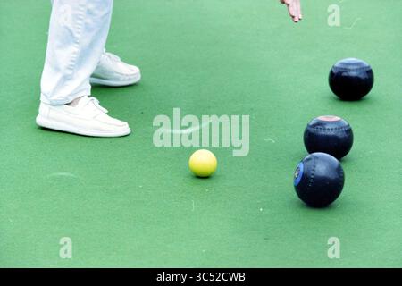 Bowls Ladies: Netherlands - England, 26-06-1999 Whizgle News, Dutch Desk, Niederlande, 1950-2000 Eine Person in weißen Schuhen positioniert sich über einem leuchtend gelben Ball auf einer grünen Oberfläche, umgeben von drei dunklen Bowlen, und bereitet sich auf ihren nächsten Zug in einem Rasenbowling vor. Stockfoto