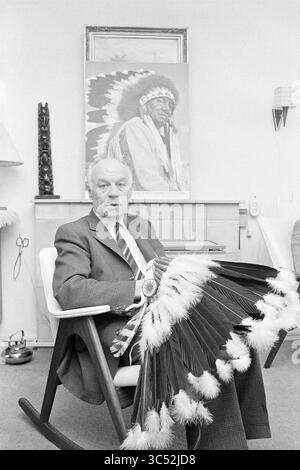 Posierender Mann, sitzend mit indischem Kopfschmuck Whizgle News, Dutch Desk, Niederlande, 1950-2000 Ein angesehener Mann sitzt auf einem modernen Stuhl, hält einen kunstvollen Federfächer, mit einem Porträt eines Häuptlings der Ureinwohner Amerikas, das hinter ihm hängt. Seine Kleidung deutet auf Formalität hin, kontrastiert mit dem kulturellen Emblem, das er hält, und schafft einen reichen Dialog zwischen Tradition und zeitgenössischem Leben. Stockfoto