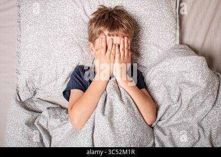 Das Kind hatte Angst vor dem Schlafengehen. Nachtschrecken bei einem Kind. Der Junge bedeckt sein Gesicht mit seinen Händen in Angst. Kindererlebnisse. Stockfoto