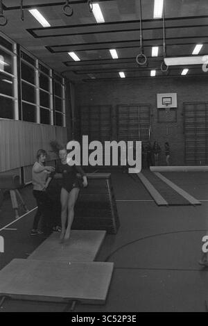 Gymnastikverband IJmuiden, Gymnastik, 15-02-1972 Whizgle News, Dutch Desk, Niederlande, 1950-2000 Eine Turnerin bereitet sich darauf vor, von einer Matte zu springen, während ein Trainer sie von der Seite unterstützt. Im Hintergrund beobachtet eine Gruppe von Zuschauern ihre Praxis in einem Indoor-Gymnasium, umgeben von Trainingsgeräten und Trainingsgeräten. Stockfoto