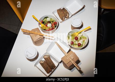 Draufsicht zeigt Speisen zum Mitnehmen auf dem Tisch, einschließlich Salate mit Tomaten und Käse, Sushi-Brötchen in Boxen, Einweg-Kaffeetassen, Essstäbchen, gelbe Plastikgabeln, keine Personen sichtbar Stockfoto