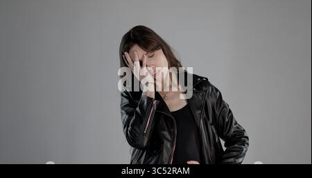 Junge Frau mit langen braunen Haaren in schwarzer Kleidung und Lederjacke, die ihr Gesicht mit einer Hand bedeckt, geschämt oder gelangweilt Stockfoto
