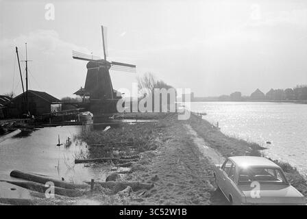 Mill 'De Eenhoorn', Mills, 06-03-1968 Whizgle News, Dutch Desk, Niederlande, 1950-2000 Eine ruhige Flusslandschaft bietet eine traditionelle Windmühle und ein ruhiges Gewässer mit Holzbooten, die an einem Dock befestigt sind. Hohe Gräser und ein gewundener Pfad führen zum fernen Horizont, wo man bezaubernde Gebäude mit Silhouetten am Himmel sehen kann. Ein Oldtimer parkt in der Nähe und verleiht der Landschaft einen nostalgischen Touch. Stockfoto