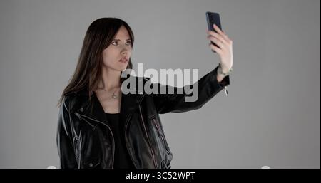 Junge Frau, die ein Selfie mit Smartphone macht, trägt eine schwarze Lederjacke auf grauem Hintergrund Stockfoto