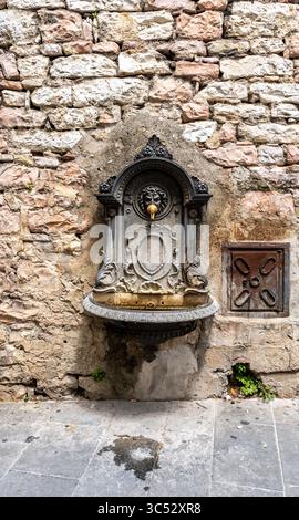 Kunstvoller öffentlicher Brunnen aus Gusseisen mit Löwenkopf an einer mittelalterlichen Steinmauer in einer historischen Straße von Assisi, Umbrien, Italien. Stockfoto