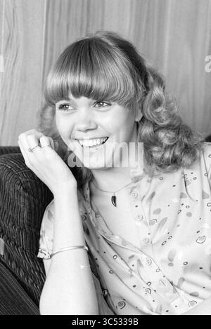Sylvia Sluyter Schauspielerin A'Dam, Dreharbeiten, Filmaufnahmen, Amsterdam, Niederlande, 28-09-1979 Whizgle News, Dutch Desk, Niederlande, 1950-2000 Eine junge Frau mit lockigen Haaren und einem fröhlichen Lächeln legt ihr Kinn auf die Hand und strahlt Wärme und Charme aus. Sie trägt eine gemusterte Bluse und scheint einen Moment des Lachens zu genießen, umgeben von einer gemütlichen Umgebung. Stockfoto