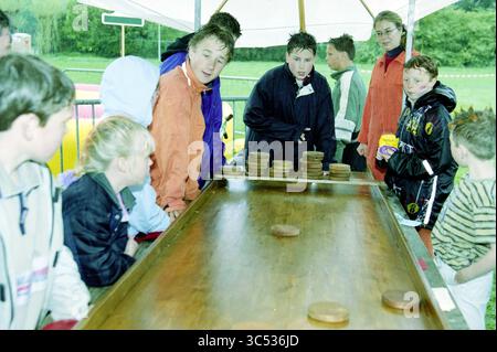 Tag der Königin im Regen mit Gesichtsbemalung, Shuffleboard und Flohmarkt in Bennebroek., Bennebroek, 04.04.2003 Whizgle News, Dutch Desk, Niederlande, 30 1950-2000 Eine Gruppe von Kindern und Jugendlichen versammelt sich um einen hölzernen Spieltisch, konzentriert sich auf ein Wettkampf-Spiel, während einige Erwachsene in der Nähe zusehen. Die Atmosphäre ist lebhaft und voller Vorfreude, während die Spieler ihre nächsten Schritte planen. Stockfoto