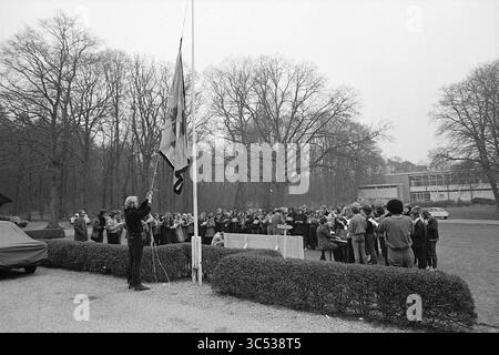 Aktionen bei CIOS Sporttraining bei Duinlust Whizgle News, Dutch Desk, Niederlande, 1950-2000 Eine Gruppe von Menschen versammelt sich draußen in der Nähe eines Fahnenmastes, während eine Person die Flagge hebt. Bäume säumen den Hintergrund, und die Szene vermittelt ein Gefühl von Festlichkeit und Gemeindeversammlungen. Stockfoto