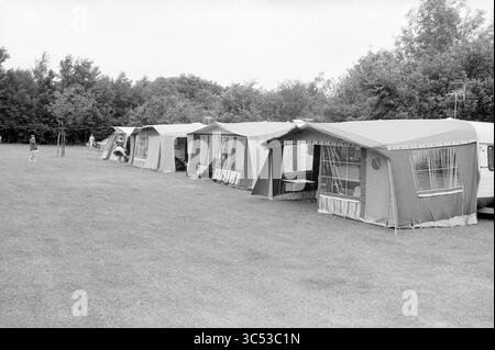 Tag Campingplatz Beeckestijn, Camping, Campingplätze, Campingplätze, Campingplatz, Campingplatz, Camping, 04-08-1983 Whizgle News, Dutch Desk, Niederlande, 1950-2000 Eine Reihe von Zelten auf einem grasbewachsenen Feld, umgeben von Bäumen. Jedes Zelt verfügt über eine Markise und einen Vordereingang, was eine gemütliche Atmosphäre auf dem Campingplatz schafft. Im Hintergrund sieht man eine Figur, die gemütlich schlendert. Stockfoto
