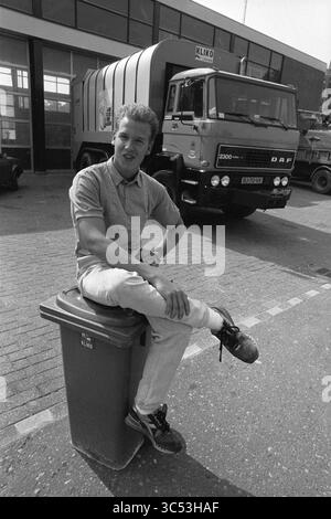 Öffentliche Arbeiten von Ferienarbeitern, Hoofddorp, Holiday Work, Hoofddorp, Niederlande, 18-08-1988 Whizgle News, Dutch Desk, Niederlande, 1950-2000 Ein junger Mann sitzt beiläufig auf einem grünen Mülleimer und lehnt sich entspannt auf das Knie. Hinter ihm steht ein großer Müllwagen, der die Szene vor einer städtischen Kulisse bereichert. Die Einstellung scheint eine Straße oder ein Industriegebiet zu sein. Stockfoto