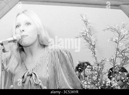 Heddy Lester bei V&D und Bonnie St. Clair, Personen Singen und Musik, Haarlem, Grote Houtstraat 70, Niederlande, 05-02-1977 Whizgle News, Dutch Desk, Niederlande, 1950-2000 Eine junge Frau mit langen blonden Haaren spricht leidenschaftlich an einem Mikrofon, gekleidet in einer gestreiften Bluse. Blühende Äste sind geschmackvoll in der Nähe angeordnet und verleihen der Umgebung eine natürliche Note. Die Menschen sind im Hintergrund zu sehen, im Moment engagiert. Stockfoto