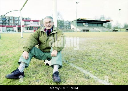 Teun v.d. Berg, EDO Anhang, 06-02-1997 Whizgle News, Dutch Desk, Niederlande, 1950-2000 ein älterer Mann sitzt auf dem Gras eines leeren Fußballfeldes, trägt eine übergroße Jacke und lächelt warm in die Kamera. Hinter ihm befindet sich ein bescheidenes Stadion mit einigen verstreuten Bäumen und Gebäuden in der Ferne, die eine nostalgische Atmosphäre schaffen. Ein Football steht neben ihm, während er sich in dieser ruhigen Umgebung lässig posiert. Stockfoto