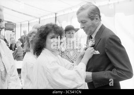 Eröffnung der Dampfpumpenstation auf halbem Weg durch Prince Claus, Eröffnung, Eröffnung, Inbetriebnahme, Dampfpumpenstation und Dampfpumpenstation, Halfweg, 10-06-1987 Whizgle News, Dutch Desk, Niederlande, 1950-2000 Eine junge Frau steckt einem elegant gekleideten Mann eine Blume zart an das Revers, der scheinbar in Gesprächen verwickelt ist. Hinter ihnen versammelt sich eine Gruppe von Menschen, darunter eine Frau mit Hut und andere in formeller Kleidung, um eine festliche Atmosphäre zu schaffen. Stockfoto