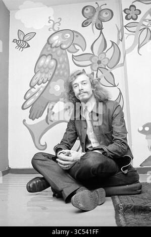 Heer posiert vor seiner Kreation Whizgle News, Dutch Desk, Niederlande, 1950-2000 Ein junger Mann mit langen, lockigen Haaren sitzt im Kreuz auf dem Boden, trägt eine Jeansjacke und eine entspannte Hose. Hinter ihm befindet sich ein lebhaftes Wandbild mit einem Papagei und skurrilen Blumen, die der Szene eine verspielte Atmosphäre verleihen. Er wirkt nachdenklich und entspannt, während er leicht aus der Kamera blickt. Stockfoto