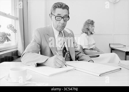 Porträt eines Mannes Whizgle News, Dutch Desk, Niederlande, 1950-2000 Ein Mann in formeller Kleidung, mit Brille, konzentriert sich darauf, Notizen in einem Notizbuch zu machen, während eine Frau im Hintergrund sitzt und leise an einem Tisch mit einer Tasse beobachtet. Weiches Licht filtert durch ein Fenster und unterstreicht die einfache, professionelle Atmosphäre. Stockfoto