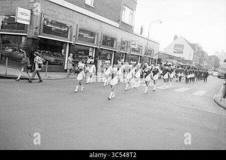 Harmonie NZH, Noord Zuid Hollandse Vervoersmaatschappij N.V., NZ, 02-07-1971 Whizgle News, Dutch Desk, Niederlande, 1950-2000 Eine farbenfrohe Parade führt durch eine belebte Straße, in der die Teilnehmer Uniformen tragen und Fahnen tragen, während sich die Zuschauer versammeln, um zu beobachten. Ladenfronten säumen die Szene, und mehrere Fahrzeuge parken in der Nähe, was zur lebhaften Atmosphäre beiträgt. Stockfoto