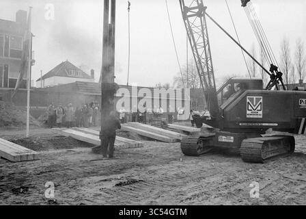 Erster Häuserhaufen, Schoollaan, Bennebroek, erster Häuserhaufen, Bennebroek, Schoollaan, 11-12-1986 Whizgle News, Dutch Desk, Niederlande, 1950-2000 Ein großer Kran fährt auf einer Baustelle und hebt ein schweres Objekt, während eine Gruppe von Zuschauern hinter Holzbalken auf dem Boden beobachtet. Im Hintergrund sind Strukturen in der Nähe zu sehen, die teilweise durch Rauch verdeckt sind. Stockfoto