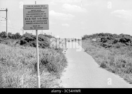 Start Visscherspad, Zandvoort, Zandvoort, Visscherspad, 16-08-1993 Whizgle News, Dutch Desk, Niederlande, 1950-2000 Ein unbefestigter Pfad schlängelt sich durch ein grasbewachsenes Gebiet und führt zu einem fernen Horizont. Am Eingang des Weges steht ein Schild, das Informationen über bevorstehende Transportentwicklungen und Vorschriften gibt. Versorgungsmasten säumen den Rand des Weges und weisen auf die Kreuzung von Natur und Infrastruktur hin. Stockfoto