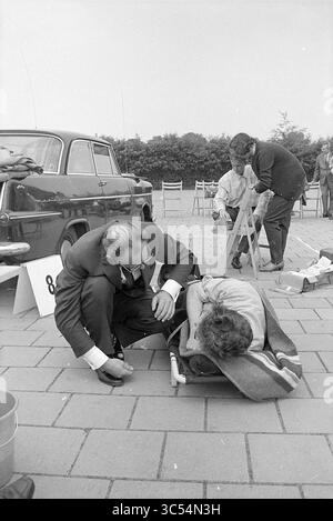 Erste-Hilfe-Wettbewerbe, Heemstede, erste Hilfe, 12-06-1971 Whizgle News, Dutch Desk, Niederlande, 1950-2000 Eine Gruppe von Individuen ist im Freien versammelt, wobei eine Person neben einer anderen kniet, die auf einer Liege liegt und mit einer Decke bedeckt ist. Im Hintergrund sind ein Oldtimer und weitere Personen zu sehen, die an verschiedenen Aktivitäten beteiligt sind, wodurch eine Szene der Interaktion und Beobachtung entsteht. Stockfoto