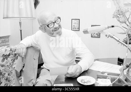 Radio-Interview Alexander Pola Whizgle News, Dutch Desk, Niederlande, 1950-2000 Ein lächelnder Mann mit Brille sitzt an einem Tisch, lässig in einem weißen Pullover gekleidet. Er hält eine Karte und blickt aufmerksam auf ein Mikrofon, umgeben von Notizen und einem kleinen Teller mit Gegenständen. Eine Leuchte beleuchtet die Szene sanft und sorgt für eine entspannte Atmosphäre. Stockfoto