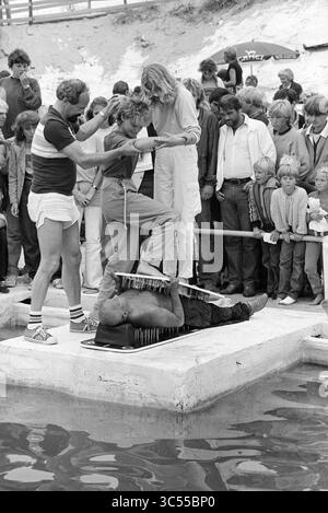 Miss Beach Election Zandvoort II, Miss Election, Zandvoort, Zandvoort, 08-08-1981 Whizgle News, Dutch Desk, Niederlande, 1950-2000 Eine Menge versammelt sich um einen Darsteller, der auf einer Plattform liegt, und demonstriert eine gewagte Leistung, bei der er ein schweres Objekt auf seiner Brust balanciert, während andere ihm helfen und sich mit ihm beschäftigen. Spannung und Neugierde sind auf den Gesichtern der Zuschauer zu sehen, wenn sie beobachten, wie sich das Spektakel am Ufer des Wassers entfaltet. Stockfoto