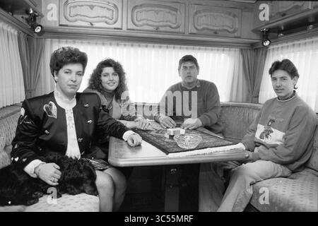 Fair Family de Nies, Haarlem, Niederlande, 01-05-1991 Whizgle News, Dutch Desk, Niederlande, 1950-2000 Eine Gruppe von vier Personen versammelt sich an einem gemütlichen Tisch in einem Mobilheim, um sich zu unterhalten. Eine Person trägt eine stilvolle Jacke, die einen schwarzen Hund sanft wiegt, während andere aufmerksam sitzen und eine warme und einladende Atmosphäre schaffen. Stockfoto