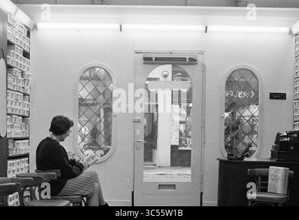 Stille in Geschäften, Geschäften und Kiosken, 12-02-1963 Whizgle News, Dutch Desk, Niederlande, 1950-2000 Eine Frau sitzt leise in einem gemütlichen Geschäft, ihre Aufmerksamkeit konzentriert sich auf ein Buch. Der Eingang verfügt über elegante Spiegelfenster, die einen Blick auf die Straße vor dem Hotel bieten. Die Atmosphäre ist ruhig und einladend, eingerahmt von Regalen mit ordentlich angeordneten Gegenständen. Stockfoto