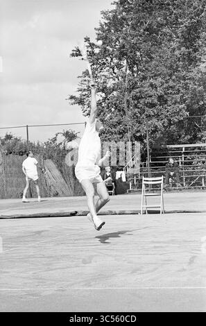 Tennis Brederode III, Jaidip Mukerjea-Tom Okker, Santpoort, 197-07-1965 Whizgle News, Dutch Desk, 30 Niederlande, 1950-2000 Ein Tennisspieler in der Luft, der den Ball mit Intensität auf einem strukturierten Platz serviert, während ein weiterer Spieler und Sitzbereich im Hintergrund sichtbar sind, eingerahmt von Laubbäumen. Stockfoto