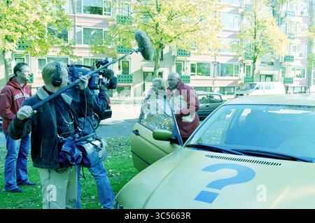 Die Postleitzahllotterie vergibt Preise in Haarlem mit Gaston Starreveld und TV-Aufnahmen davon, Haarlem, Niederlande, 22-10-2003 Whizgle News, Dutch Desk, Niederlande, 1950-2000 Eine Crew filmt draußen ein Interview, in dem ein Mann in einer roten Jacke in der Nähe eines gelben Autos mit einem Fragezeichen eine ältere Frau angreift. Ein Kamerabediener fängt den Moment ein, während ein Mikrofon positioniert ist, um sein Gespräch vor dem Hintergrund von Herbstbäumen und nahe gelegenen Gebäuden aufzunehmen. Stockfoto