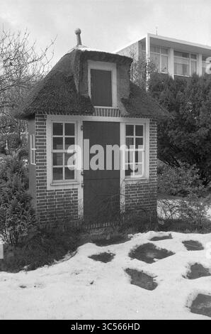 Exterieur Teahouse, Lommerlust, B'wijk, Exterieur, 26-01-1976 Whizgle News, Dutch Desk, Niederlande, 1950-2000 Ein charmantes, kleines Häuschen mit einem Strohdach steht inmitten eines bewachsenen Gartens, umgeben von Schnee- und Steinwegen, die zu seiner Holztür führen. Die skurrilen architektonischen Details und die malerischen Fenster tragen zu seinem märchenhaften Reiz bei und stehen im Kontrast zum modernen Gebäude, das im Hintergrund zu sehen ist. Stockfoto