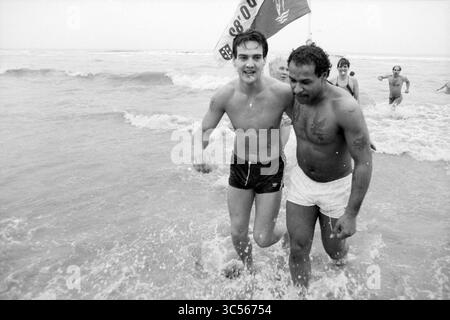 Neujahrstauchen Zandvoort, Neujahr, Zandvoort, 01-01-1987 Whizgle News, Dutch Desk, Niederlande, 1950-2000 zwei Männer laufen fröhlich durch die Wellen am Strand, lachen und genießen den Moment. Ein Mann trägt dunkle Badehosen, während der andere helle Shorts trägt. Eine Flagge im Hintergrund trägt zur Strandatmosphäre bei, und mehrere Leute können im Wasser in der Nähe spielen sehen. Stockfoto