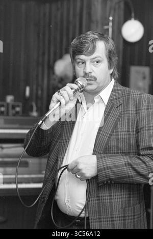 Sonstiges Künstlerfestival Zijlpoort, Gesangschöre, Haarlem, Niederlande, 24-04-1988 Whizgle News, Dutch Desk, Niederlande, 1950-2000 Ein Mann mit einem Schnurrbart hält ein Mikrofon und spricht leidenschaftlich, während er einen karierten Blazer trägt. Er steht selbstbewusst mit einem Mikrofonkabel in der Hand, was darauf hindeutet, dass er ein Publikum anspricht oder eine Vorstellung leitet. Im Hintergrund ist ein Klavier zu sehen, das auf ein musikalisches Setting hindeutet. Stockfoto