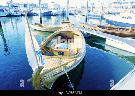HISWA J4 Holzboote, IJm., IJmuiden, Niederlande, 05-09-1996 Whizgle News, Dutch Desk, Niederlande, 1950-2000 Ein handgefertigtes Holzboot mit elegantem Design liegt an einem geschäftigen Jachthafen, umgeben von verschiedenen anderen Schiffen. Das Boot verfügt über einen markanten Bug und ein einladendes Cockpit mit einem detaillierten Innenraum, der durch seine transparente Abdeckung sichtbar ist. Das ruhige Wasser spiegelt die nahe gelegenen Docks und Gebäude wider und trägt zur ruhigen Atmosphäre des Hafens bei. Stockfoto
